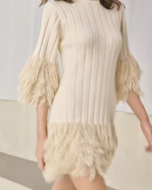 Model wearing a cream ribbed knit mini dress with textured fringe cuffs and hem walking in a minimalist studio setting.