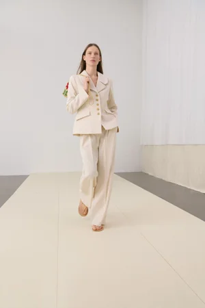Model in cream blazer and trousers walking forward against a neutral studio backdrop