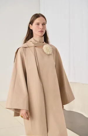 Model Angelika Wierzbicka wearing an oversized beige cape coat with a matching neck scarf and textured flower brooch for Malene Birger.