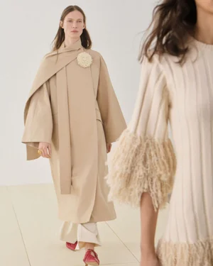 Model walking in a beige coat with draped scarf and fringed brooch for Malene Birger pre-fall lookbook.