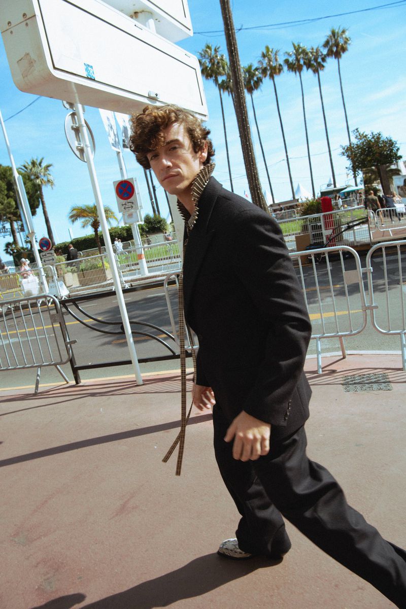Man wearing a black suit and patterned shirt walking outdoors past metal crowd control barriers and palm trees.