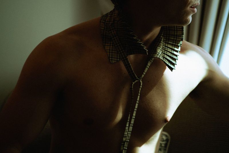 Shirtless man wearing a pleated plaid detachable collar with neck ties illuminated by directional light.