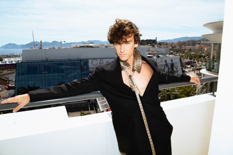 Guitarricadelafuente wearing an open black tailored jacket and a structured plaid neckpiece on a balcony overlooking the city.