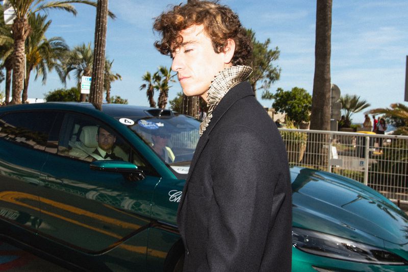 Guitarricadelafuente wearing a dark tailored coat with a ruffled collar standing outdoors beside a metallic green car.