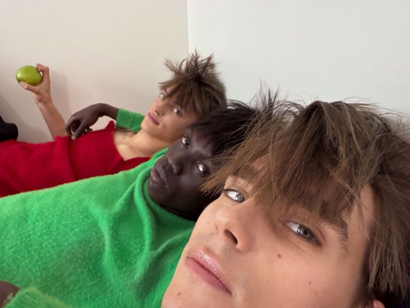 Three male models wearing green and red knitwear lying together while one holds a green apple.