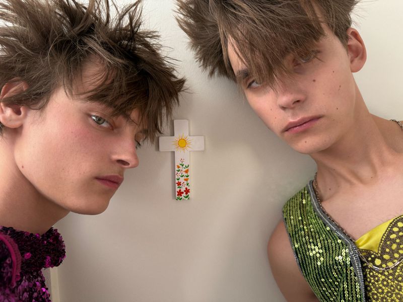 Two male models with tousled spiky hair pose next to a small floral wall cross against an off-white background.