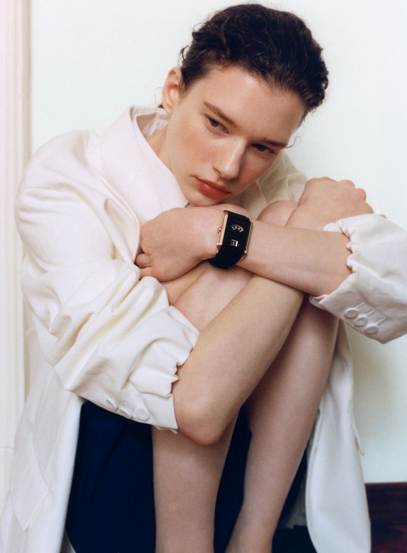 Model wearing a white jacket and a black watch, seated with knees pulled to chest against a white wall.