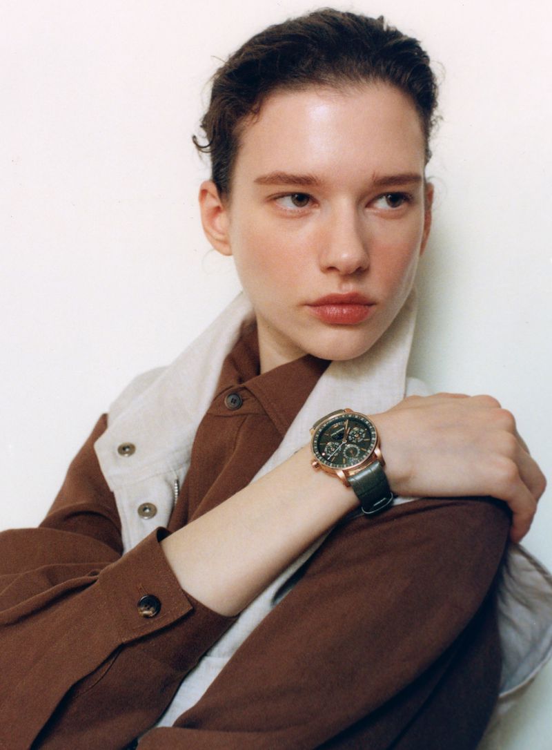 Model Sandra Murray wearing a brown button-down shirt and leather strap wristwatch against a white studio background.