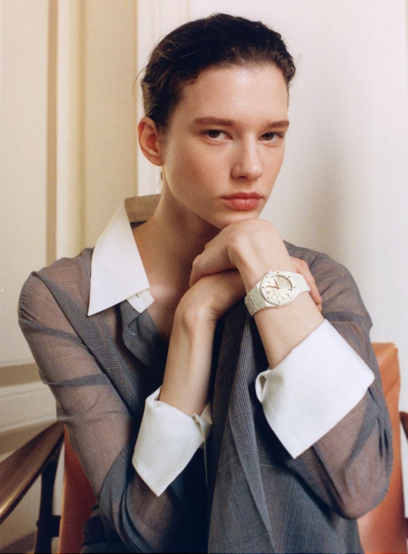 Model wearing a grey shirt with white collar and cuffs, resting chin on hands displaying a white watch against a neutral wall