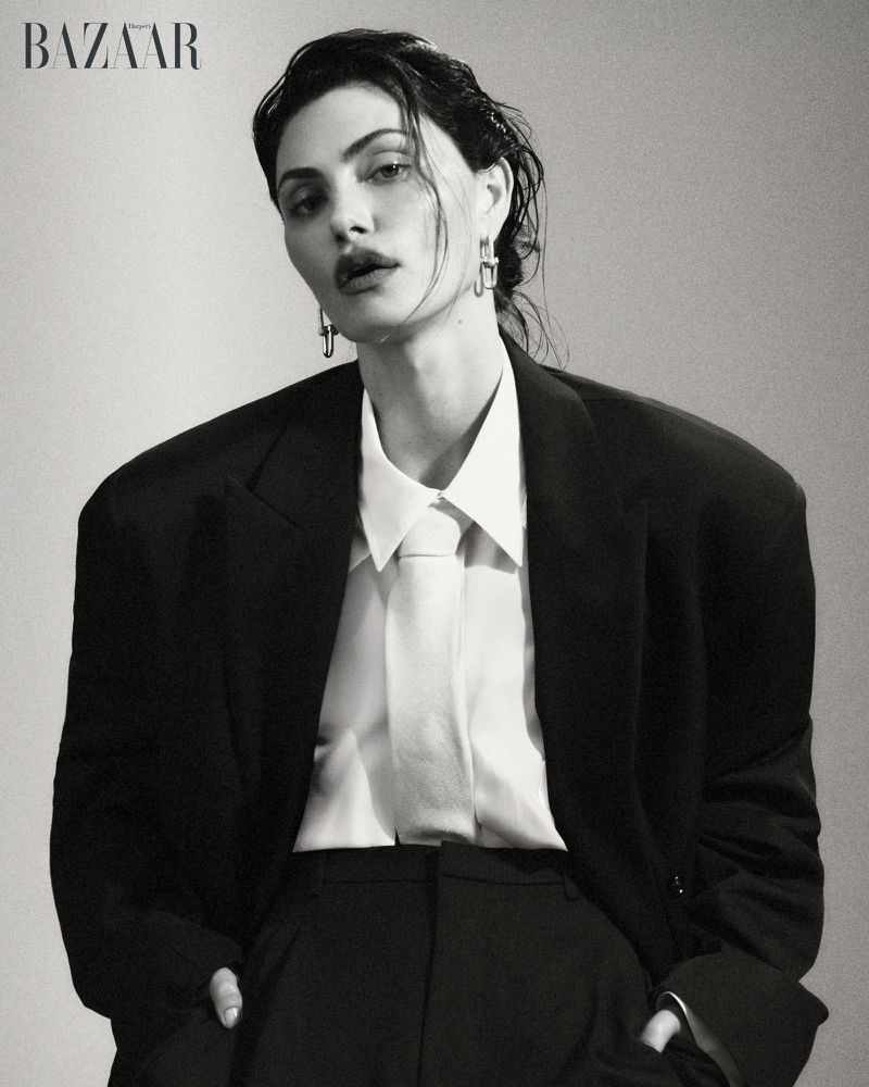 Actor Phoebe Tonkin posing in an oversized black blazer, white collared shirt, and tie against a studio backdrop for Harper's Bazaar.
