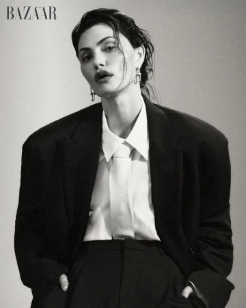 Actor Phoebe Tonkin posing in an oversized black blazer, white collared shirt, and tie against a studio backdrop for Harper's Bazaar.
