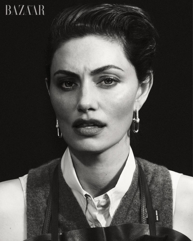 Black and white portrait of Phoebe Tonkin with slicked-back hair wearing a collared shirt, knit vest, and link earrings.