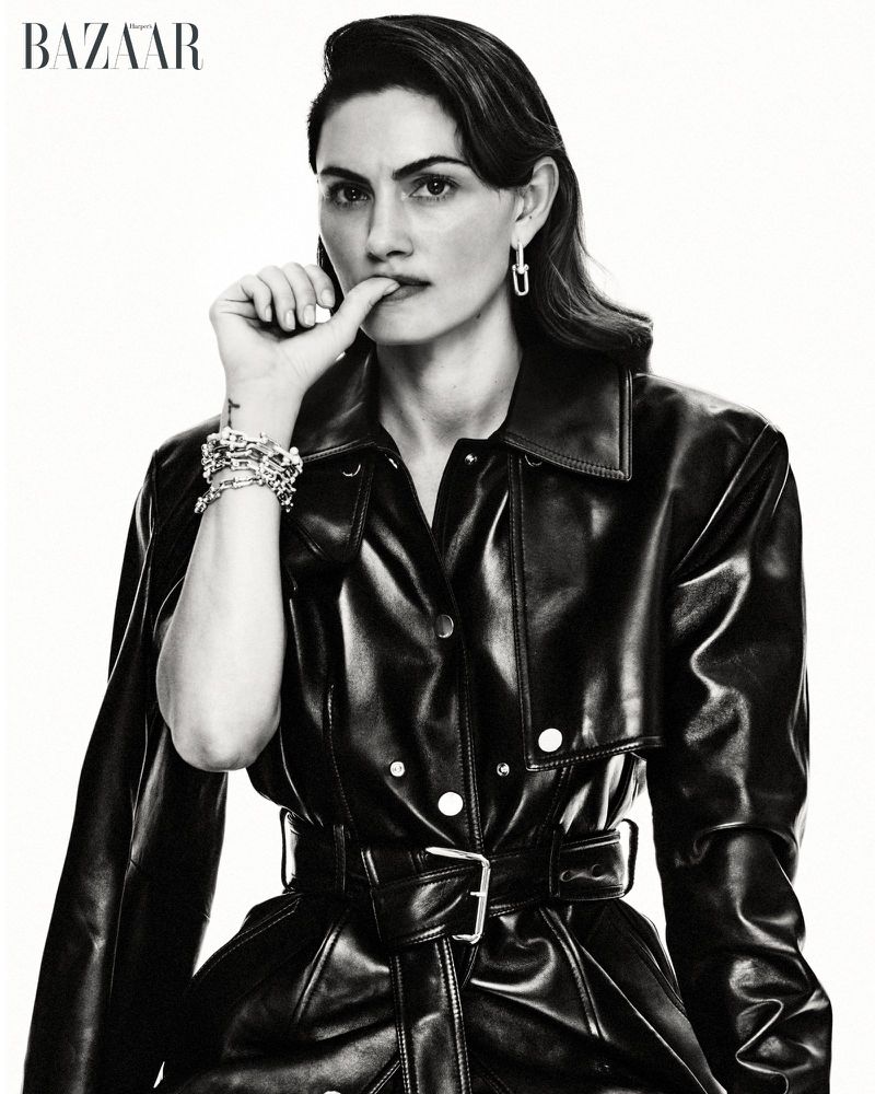Phoebe Tonkin wearing a belted black leather trench coat and silver chain jewelry against a white studio background.