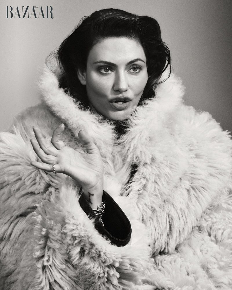 Actor Phoebe Tonkin wearing a voluminous high-collar shearling coat in a black and white editorial portrait.