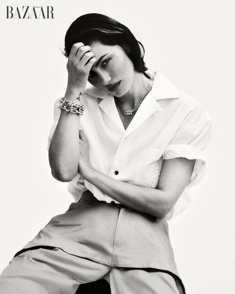 Phoebe Tonkin seated in a white button-up shirt and trousers posing with hand to head against a white background.