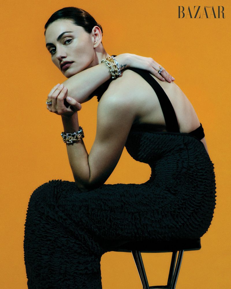 Phoebe Tonkin seated in a black textured dress with silver bracelets against an orange background for Harper's Bazaar Australia.