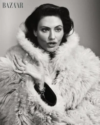 Actor Phoebe Tonkin wearing a voluminous high-collar shearling coat in a black and white editorial portrait.