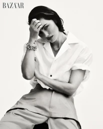 Phoebe Tonkin seated in a white button-up shirt and trousers posing with hand to head against a white background.