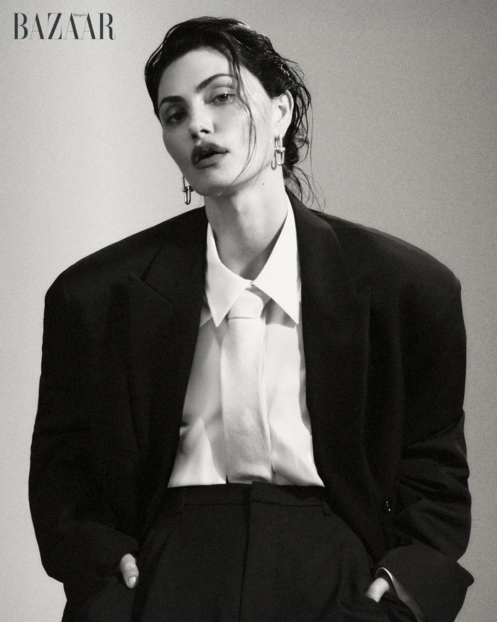 Actor Phoebe Tonkin posing in an oversized black blazer, white collared shirt, and tie against a studio backdrop for Harper's Bazaar.