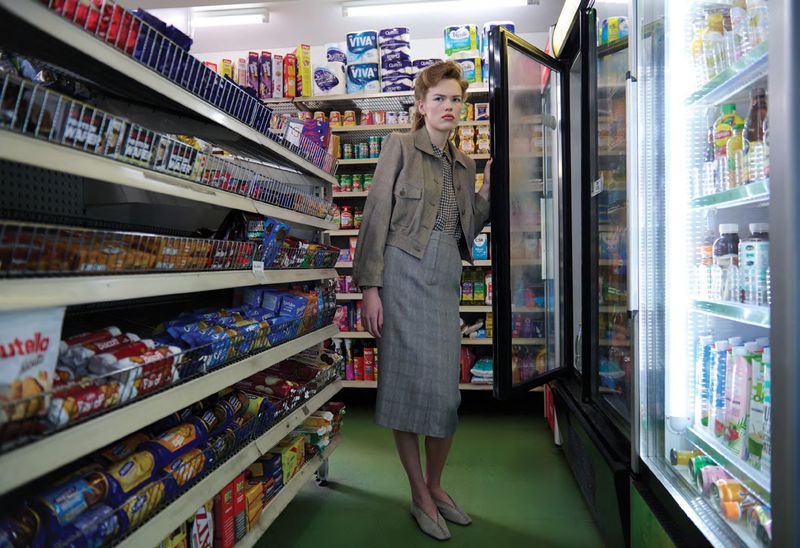 Model standing in a supermarket aisle holding open a refrigerated glass door, wearing a jacket and skirt