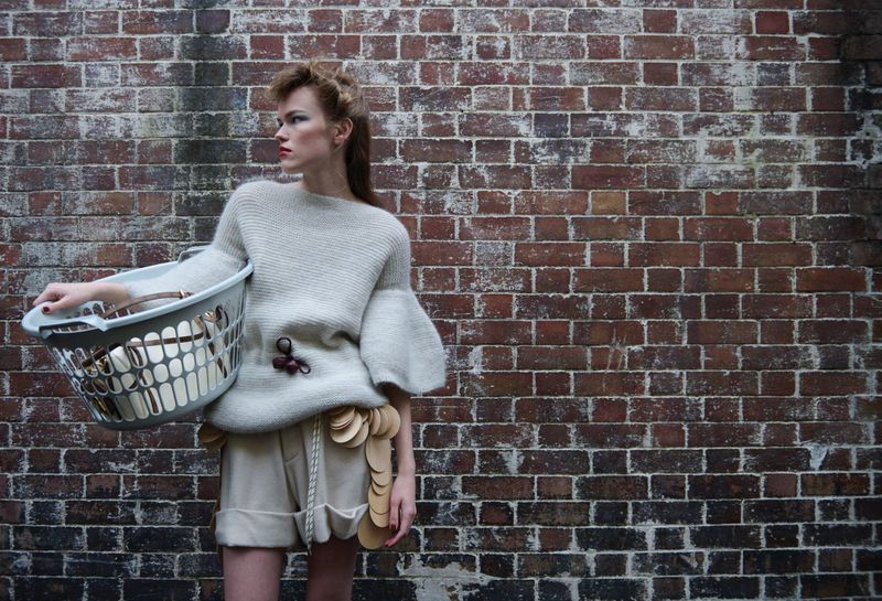 Model Mathilde Lakjaer poses against an aged brick wall holding a plastic laundry basket in an oversized knit sweater and shorts.