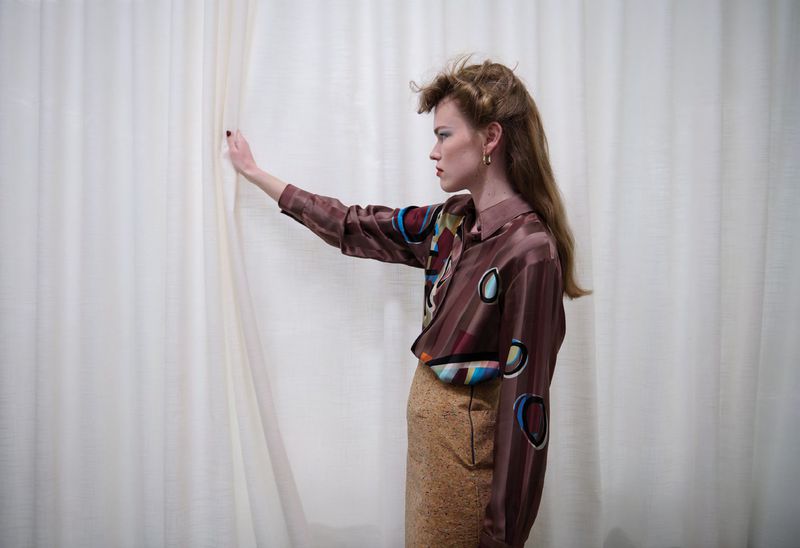 Model Mathilde Lakjaer in a printed satin blouse and textured skirt posing in profile beside a draped white curtain.