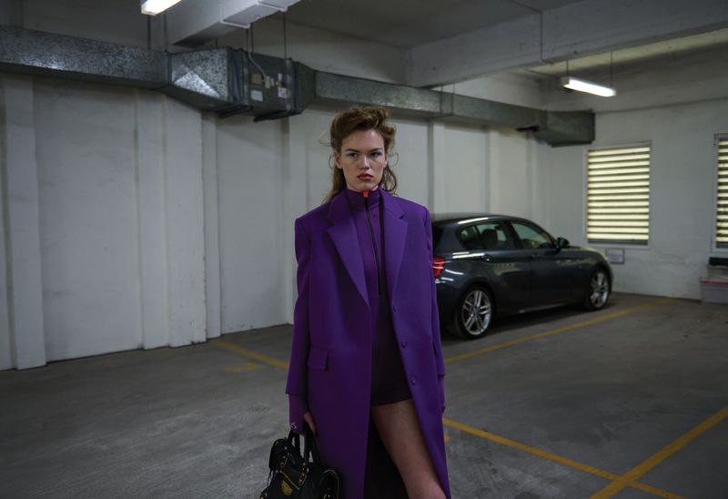 Model Mathilde Lakjaer wearing an oversized purple tailored coat and holding a black handbag in an indoor parking garage.
