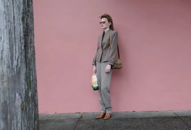Model Mathilde Lakjaer standing by a pink wall in a tailored blazer, grey trousers, sunglasses, and carrying a shoulder bag.