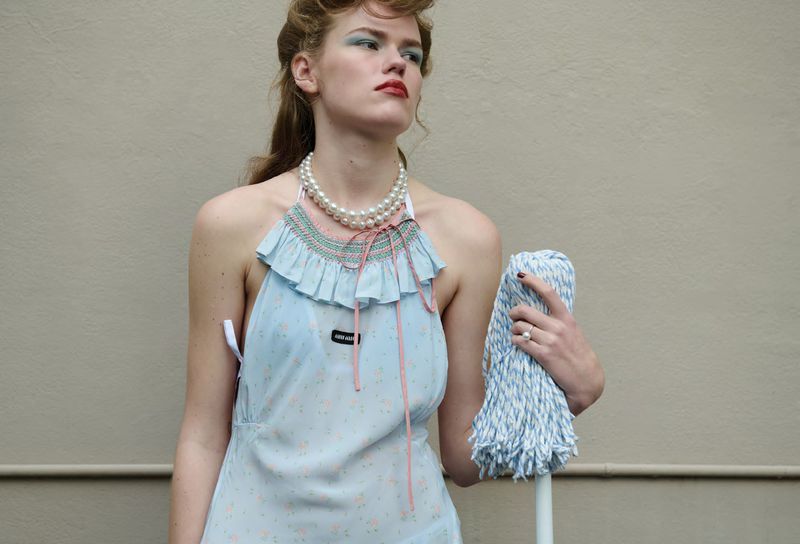 Mathilde Lakjaer wearing a light blue ruffled halter dress and pearl necklace while holding a mop against a neutral wall.
