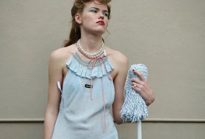 Mathilde Lakjaer wearing a light blue ruffled halter dress and pearl necklace while holding a mop against a neutral wall.