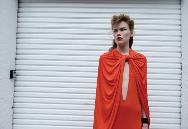 Mathilde Lakjaer wearing a draped orange cape dress in front of a white metal roller shutter.