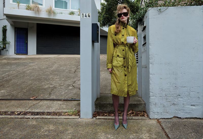 Mathilde Lakjaer in a yellow belted trench coat and sunglasses holding a white mug on a residential driveway.