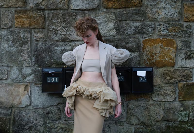 Model Mathilde Lakjaer posing in neutral tailored blazer and ruffled skirt against an exterior stone wall with black mailboxes.