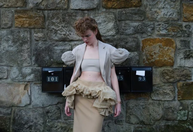 Model Mathilde Lakjaer posing in neutral tailored blazer and ruffled skirt against an exterior stone wall with black mailboxes.