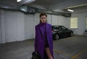 Model Mathilde Lakjaer wearing an oversized purple tailored coat and holding a black handbag in an indoor parking garage.