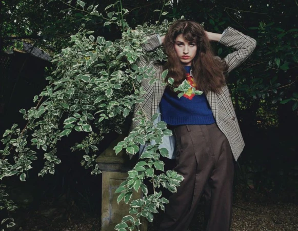 Model posing with hands behind head wearing a plaid blazer, cropped blue sweater, and brown pleated trousers among lush garden foliage.