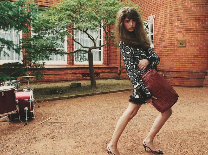Model with voluminous hair in a patterned mini dress and slingback heels holding a burgundy textured bag by an outdoor drum kit.