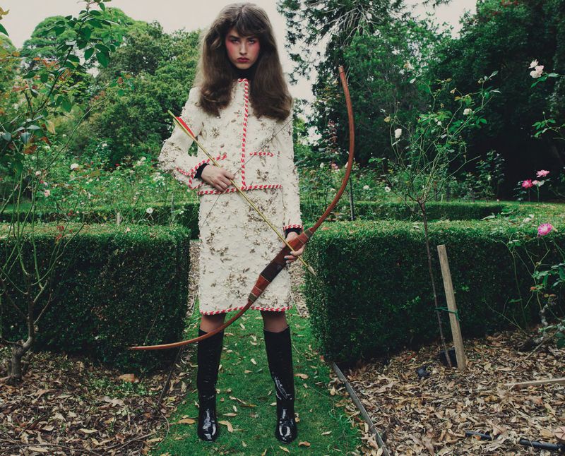 Model in a cream tweed skirt suit and black boots holding an archery bow and arrow in a hedge garden.
