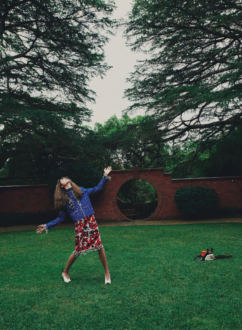 Woman posing with arms outstretched on a lawn wearing a blue jacket and floral skirt near a brick wall and chainsaw.