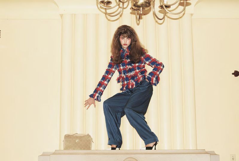 Woman in a red and blue fringed tweed jacket and wide-leg navy pants posing dynamically on a fireplace mantel beneath a chandelier.