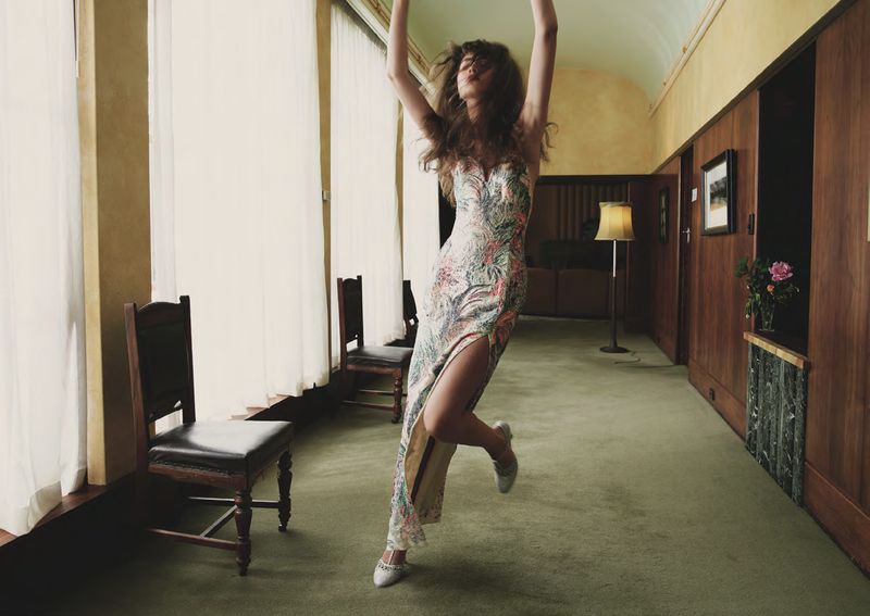 Model in a floral print slip dress dancing in a wood-paneled hallway with large windows and chairs