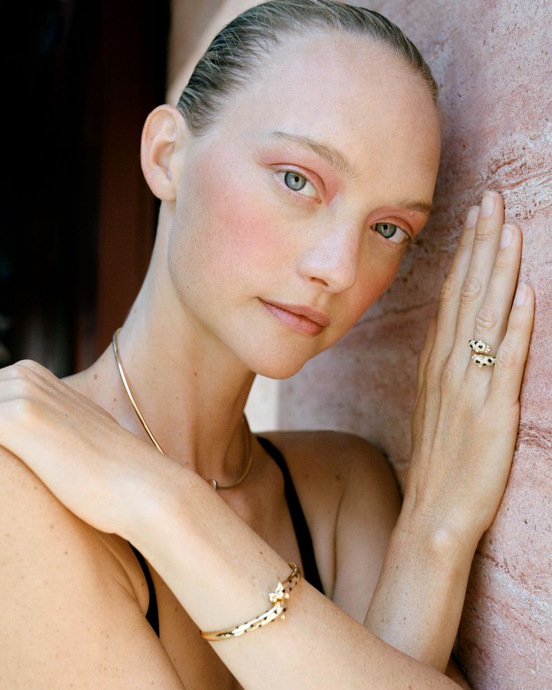 Model Gemma Ward rests her hand against a textured pink stone wall wearing fine gold jewelry and soft pink makeup.