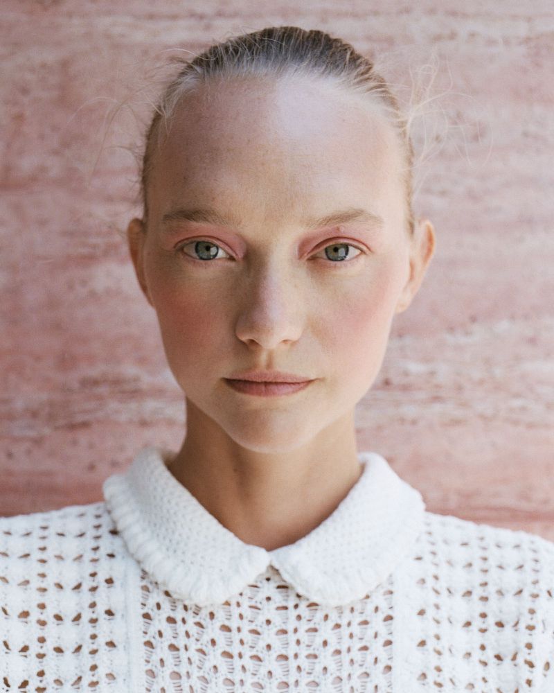 Close-up portrait of model Gemma Ward wearing a white crochet collared top against a pink textured stone backdrop.