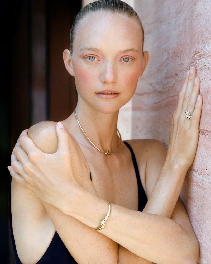 Close-up portrait of model Gemma Ward wearing gold jewelry against a warm textured stone wall.