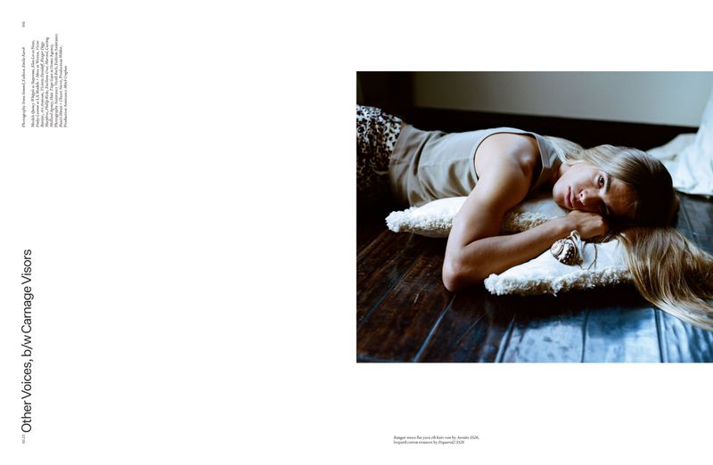 Model with long blonde hair resting on white fringe pillows over a dark wood floor for an Arena Homme + editorial.