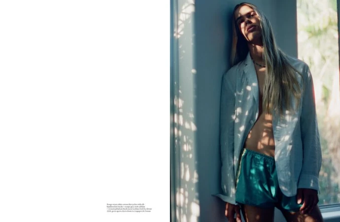 A long-haired male model stands in an open light blue jacket and green satin shorts beside a sunlit window with leafy shadows.