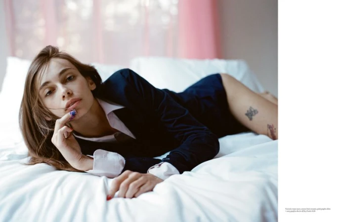 A female model reclines on a white bed wearing a dark navy button-up knit sweater and a white collared shirt.