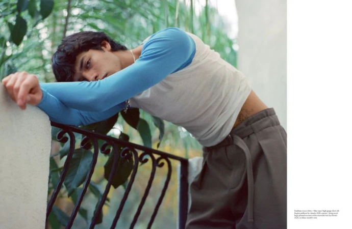 Young man wearing a blue and white long-sleeved top and grey pleated trousers leaning over an outdoor balcony railing.