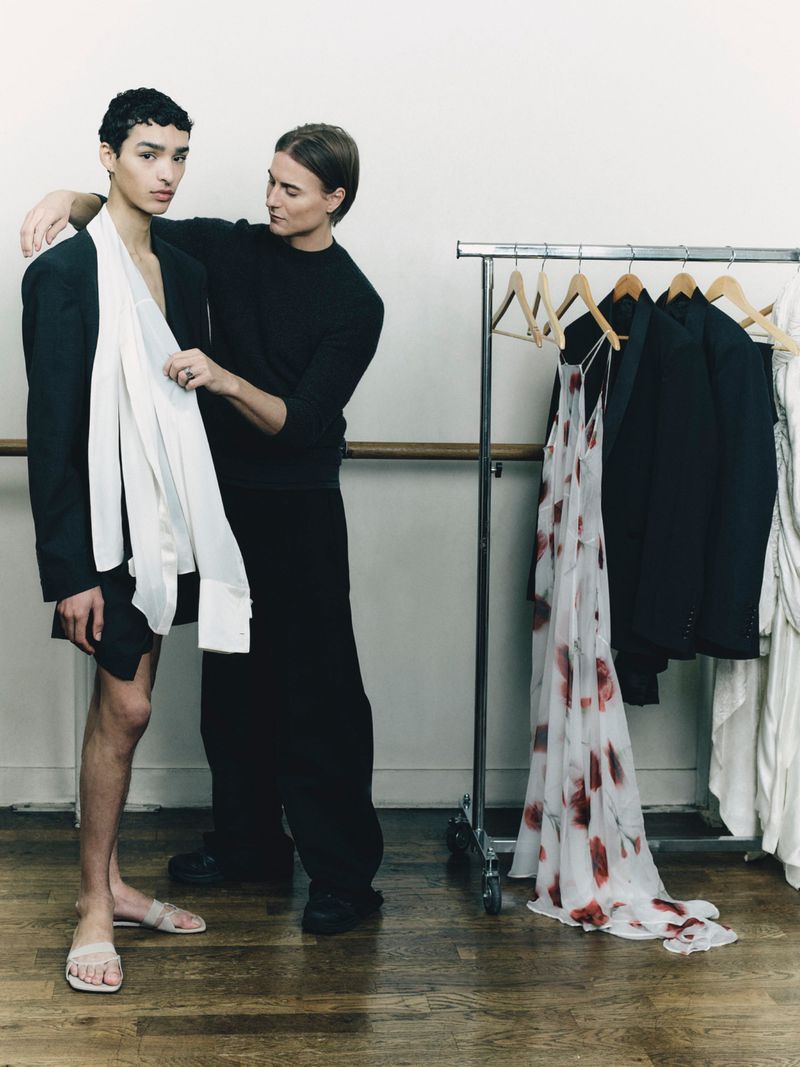 Two men posing beside a clothing rack in a studio with wooden floors, one adjusting white fabric on the other.