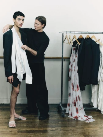 Two men posing beside a clothing rack in a studio with wooden floors, one adjusting white fabric on the other.